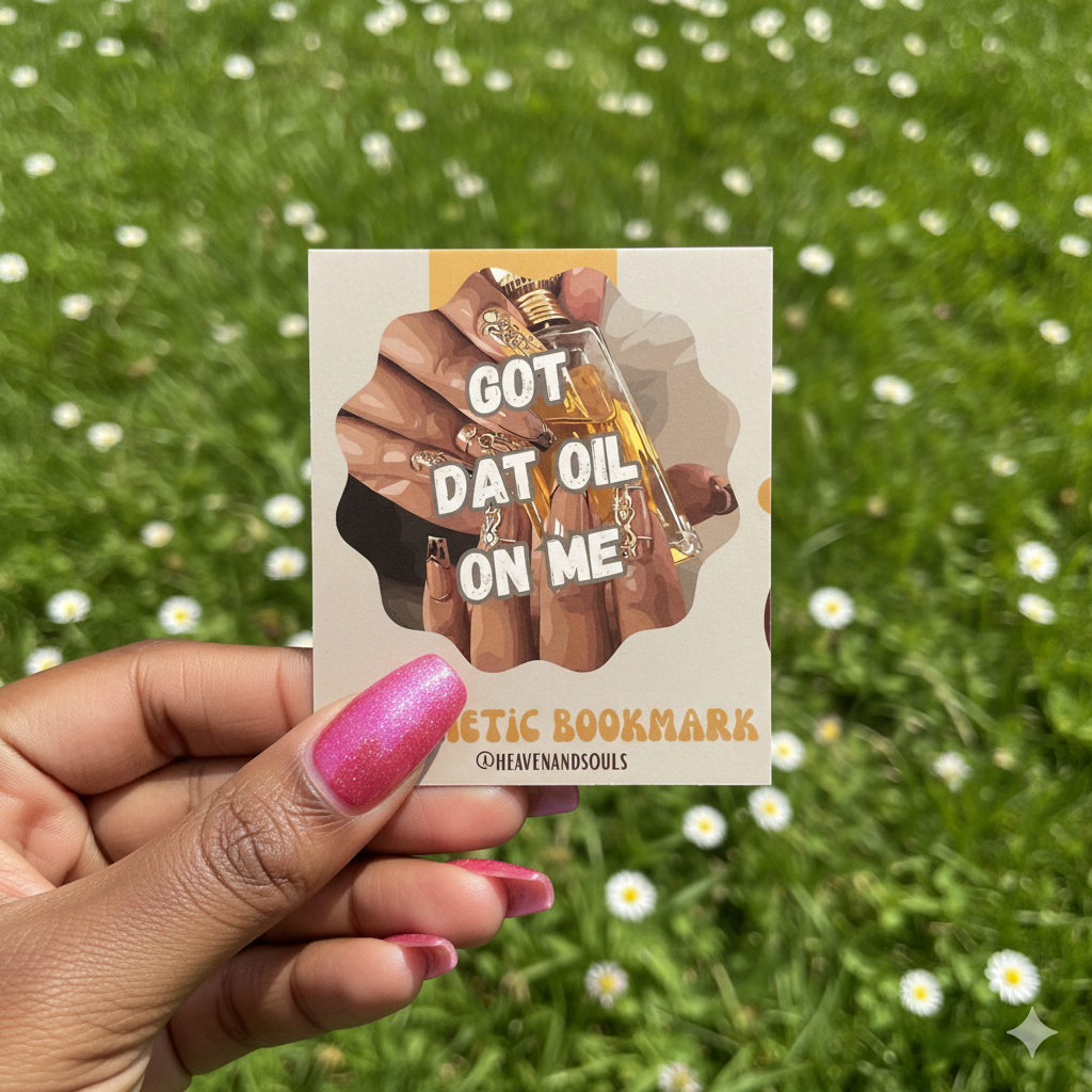 Hand holding a bookmark with a design of hands holding a bottle labeled 'GOT DAT OIL ON ME' against a grassy background.