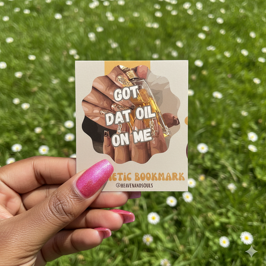 Hand holding a bookmark with a design of hands holding a bottle labeled 'GOT DAT OIL ON ME' against a grassy background.
