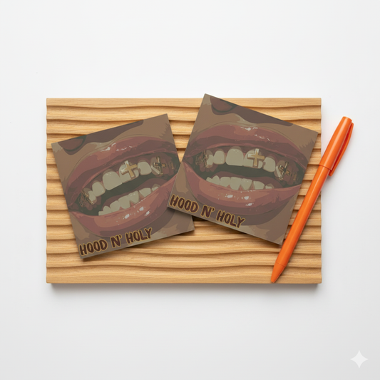 Two 'Hood N' Holy' coasters with a mouth design on a wooden surface with an orange pen.